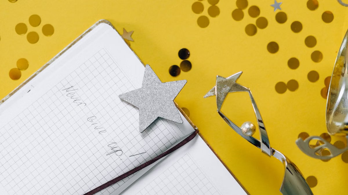 Flat lay of a notebook with motivational text, trophy, and star confetti on a yellow background.
